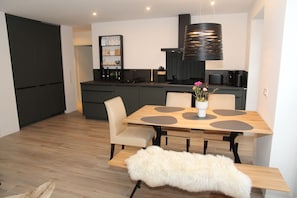 A modern kitchen with dark cabinets, a central island, and a dining area with a wooden table and chairs. - Zweite Heimat - Hochwertige Neubauwohnung Direkt am Wasser! (Krummhörn)