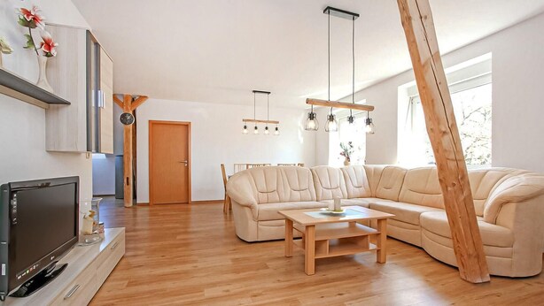 A modern living room with a large L-shaped sofa, a wooden coffee table, and a flat-screen TV on a wooden stand. There is a dining area with a wooden table and chairs, and a kitchen area with a wooden cabinet and a clock. - Ferienwohnung Zwiedorf (Zwiedorf)