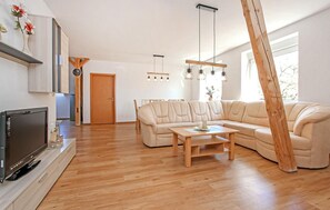 Een moderne woonkamer met een grote L-vormige bank, een houten salontafel en een flatscreen televisie op een houten dressoir. Er is een eethoek met een houten tafel en stoelen, en een keukenhoek met een houten kast en een klok.