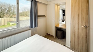 A bedroom with a bed, a window with curtains, a radiator, a wooden door, and a small trash bin. - 3 bedroom accommodation in Sandgreen (Sandgreen)