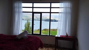 A room with a bed, a small table, and a window with a view of a lake and hills. - CABAÑAS VILLAS DEL LAGO (Cuitiva)