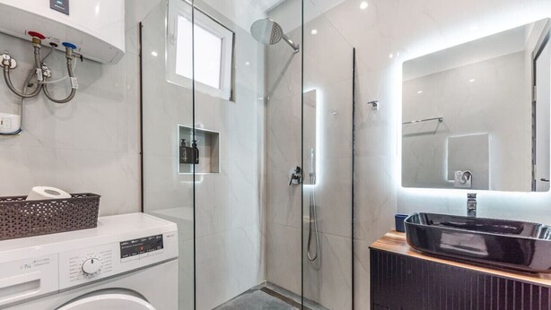 Een moderne badkamer met een douche met glazen wand, een wasmachine en een wastafel met spiegel.