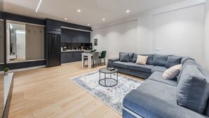 Una sala de estar moderna con un sofá gris, una mesa de centro y una zona de cocina con armarios oscuros y un refrigerador.