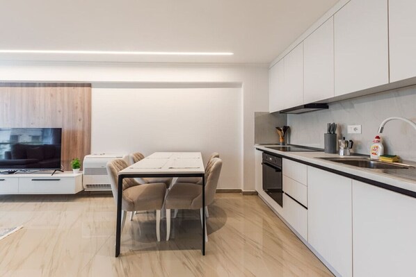 A modern kitchen with white cabinets, a black countertop, and a built-in oven. There is a dining area with a white table and chairs. - Kondo Stays - Mumba Apartment 1 (Tiranë)