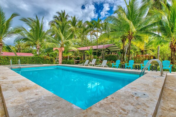 A swimming pool with clear blue water, surrounded by palm trees and a covered seating area with chairs.