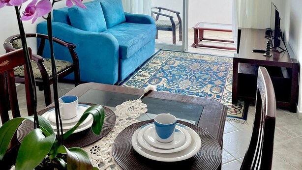 Un salon moderne avec un canapé bleu, une table à manger avec des assiettes et des tasses, un meuble pour la télévision et un balcon avec des rideaux.