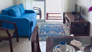 Un salon avec un canapé bleu, une petite table sur laquelle se trouvent une tasse et une soucoupe, un téléviseur posé sur un meuble en bois, et une vue à travers une porte vitrée.