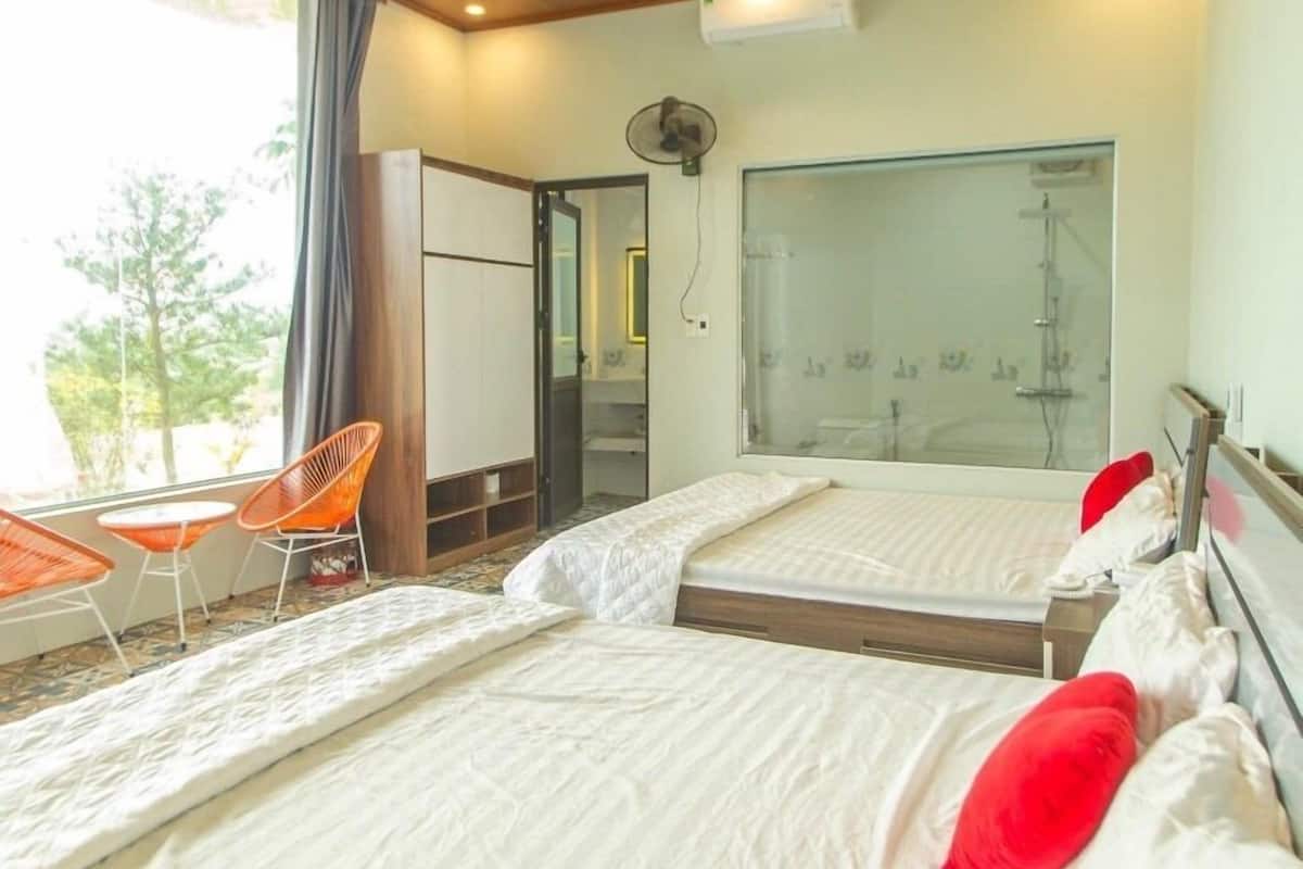 Habitación de hotel con dos camas, un ventanal grande, un balcón con sillas y zona de ducha.