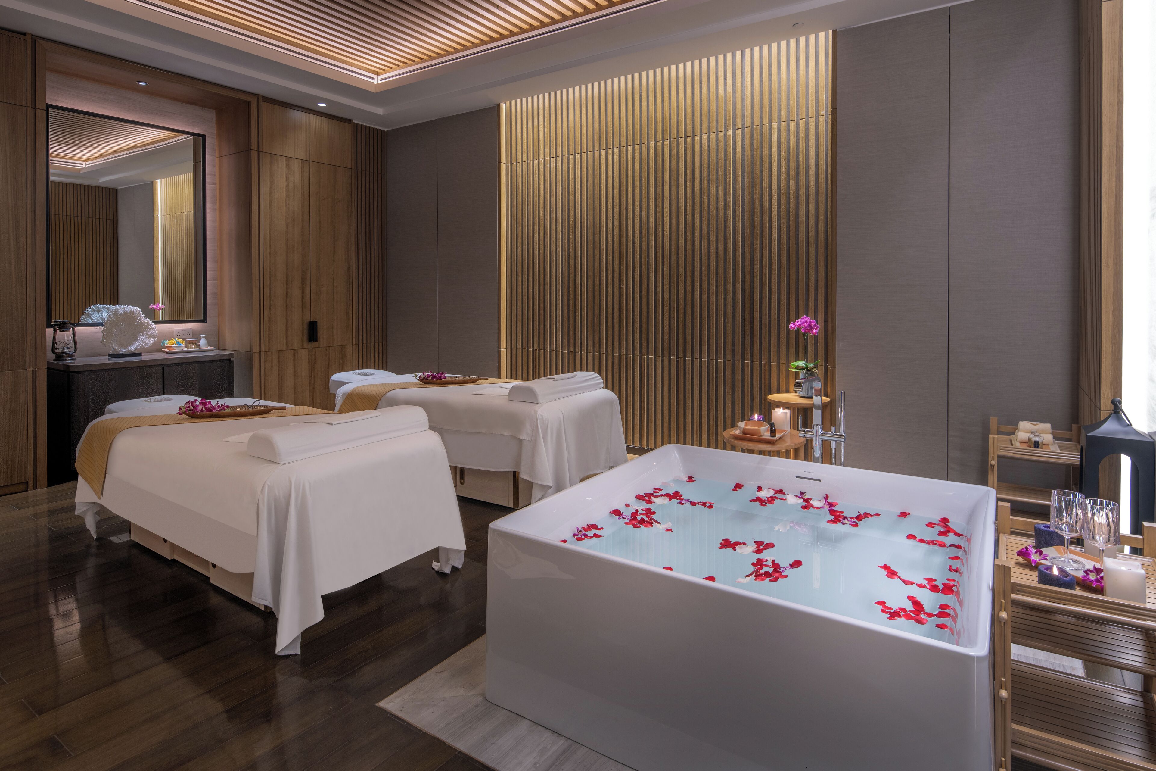 A modern spa room with a large bathtub, two massage tables, and a wooden panel wall.