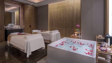 A modern spa room with a large bathtub, two massage tables, and a wooden panel wall.