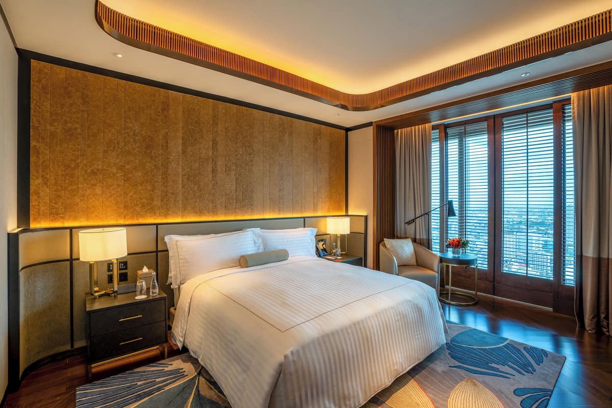 A modern hotel room with a large bed, bedside tables, a nightstand, a chair, and a large window with blinds.