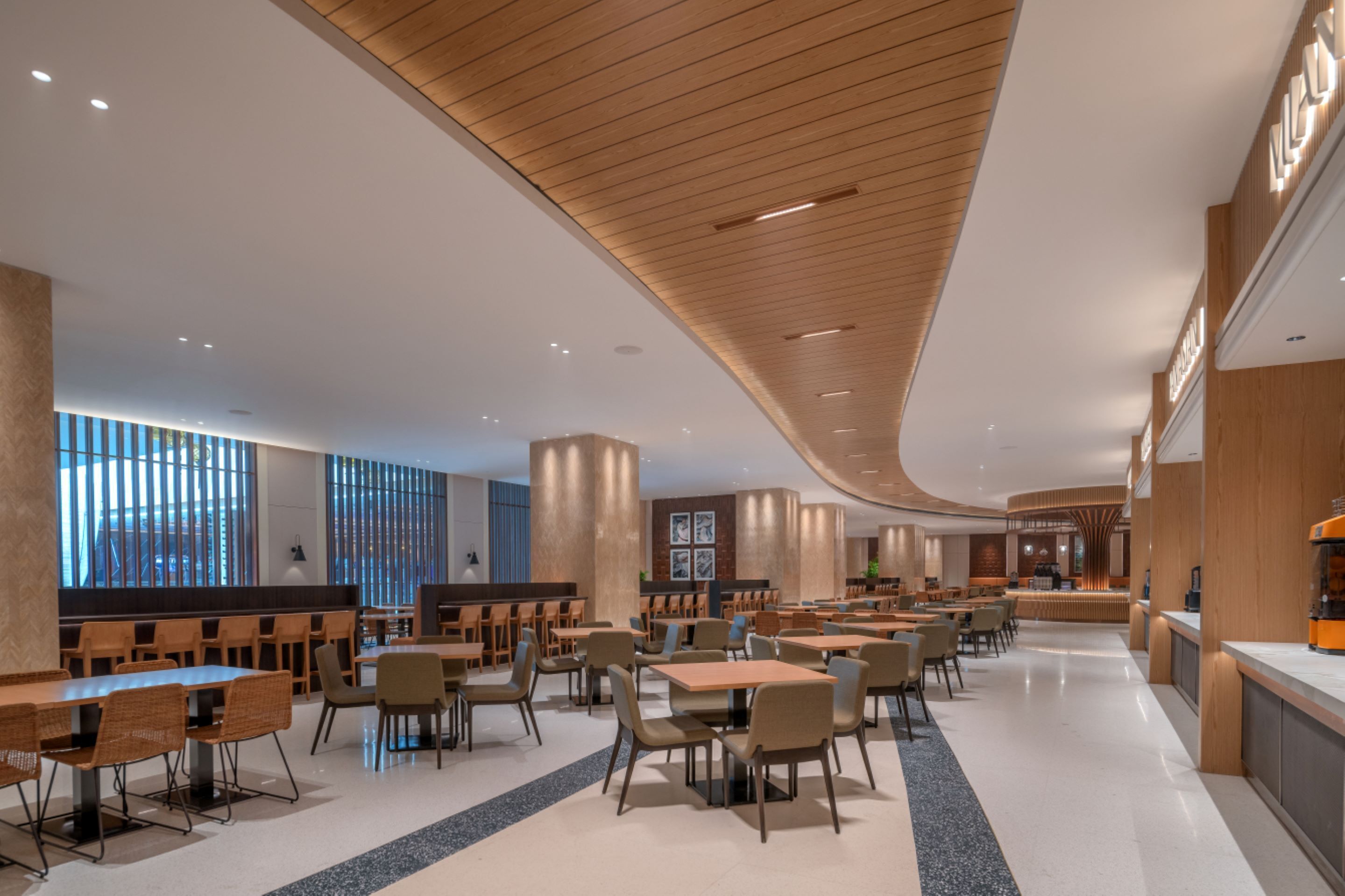 A spacious, modern dining area with multiple tables and chairs, wooden accents on the ceiling, and large windows providing natural light.