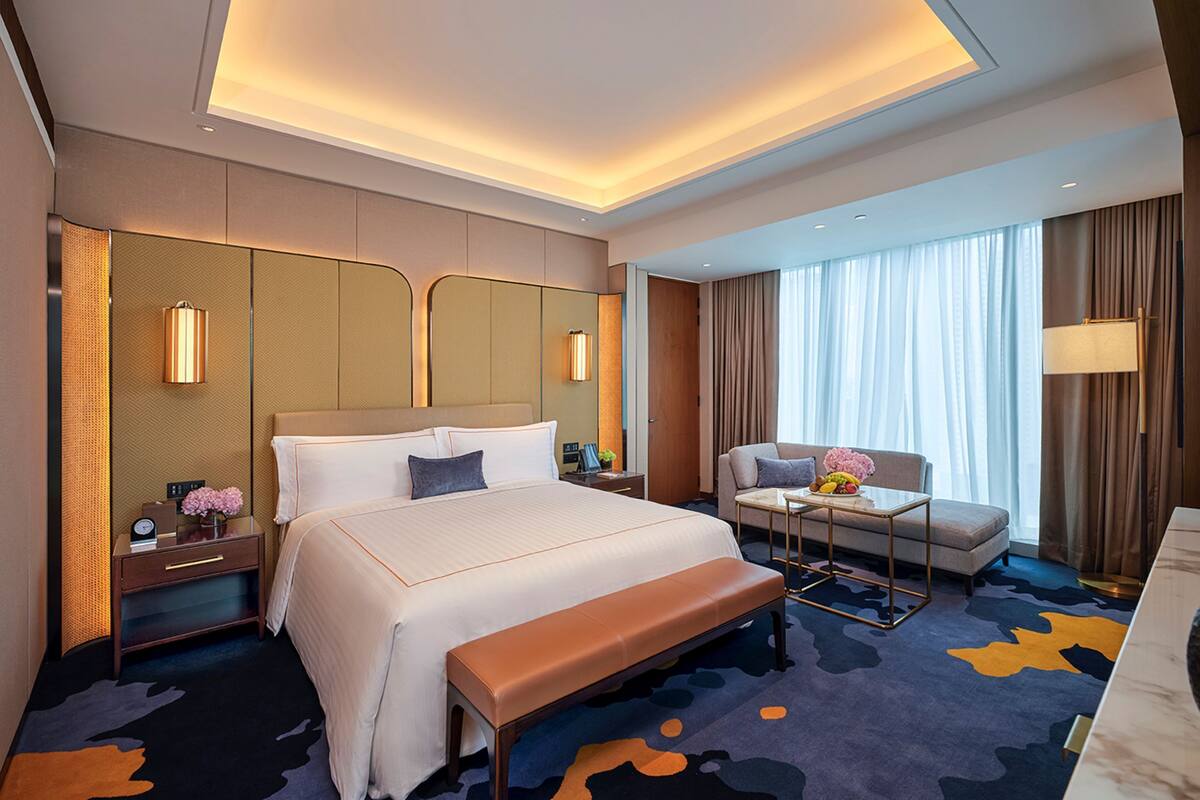 A modern hotel room with a large bed, bedside tables, a bench, a sofa, and a desk with a lamp.