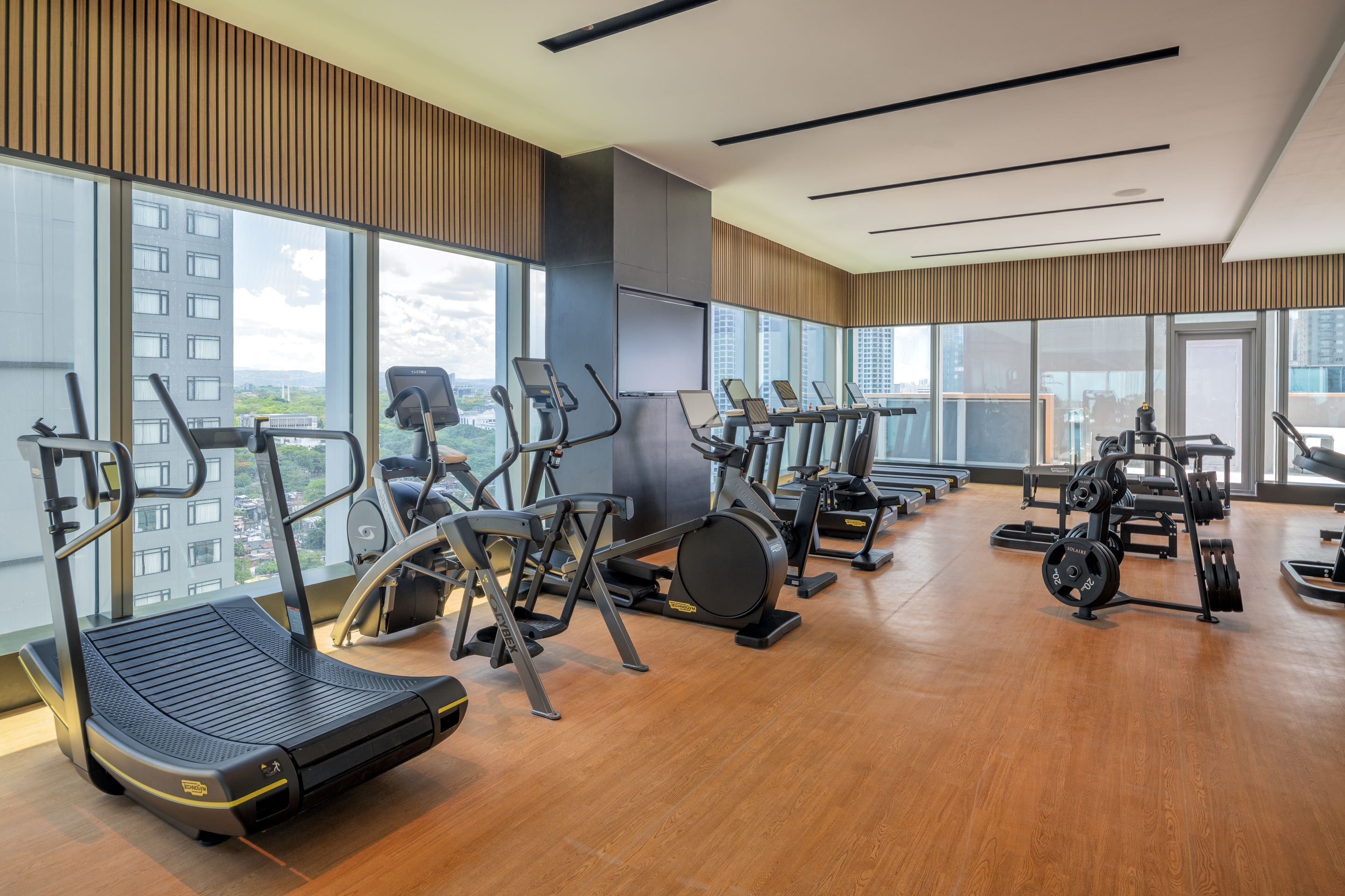 A modern gym with various exercise equipment, large windows, and a view of the cityscape.