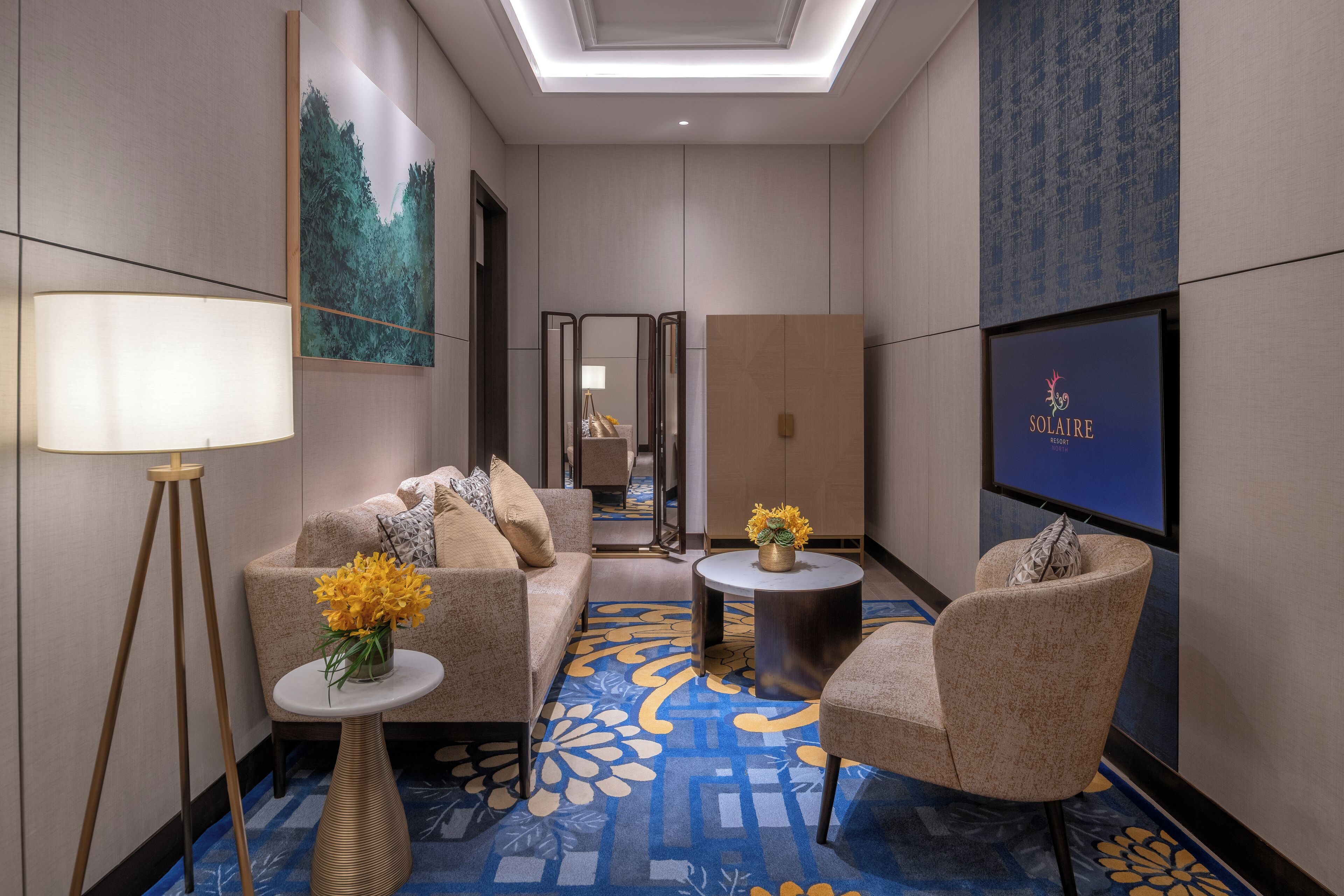 A modern hotel room with a sofa, armchairs, a small table, a floor lamp, and a flat-screen TV displaying 'SOLAIRE'.
