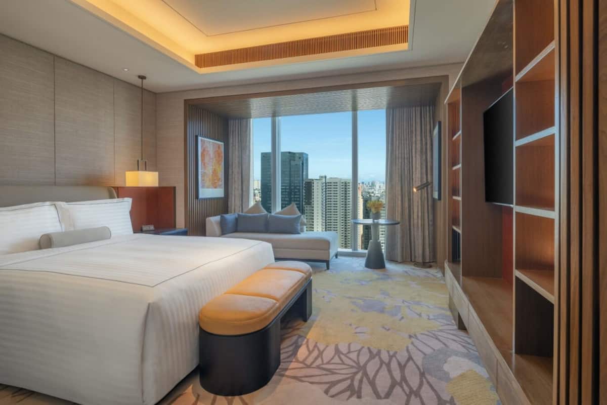 A modern hotel room with a large bed, a sofa, a TV, and a view of the cityscape.