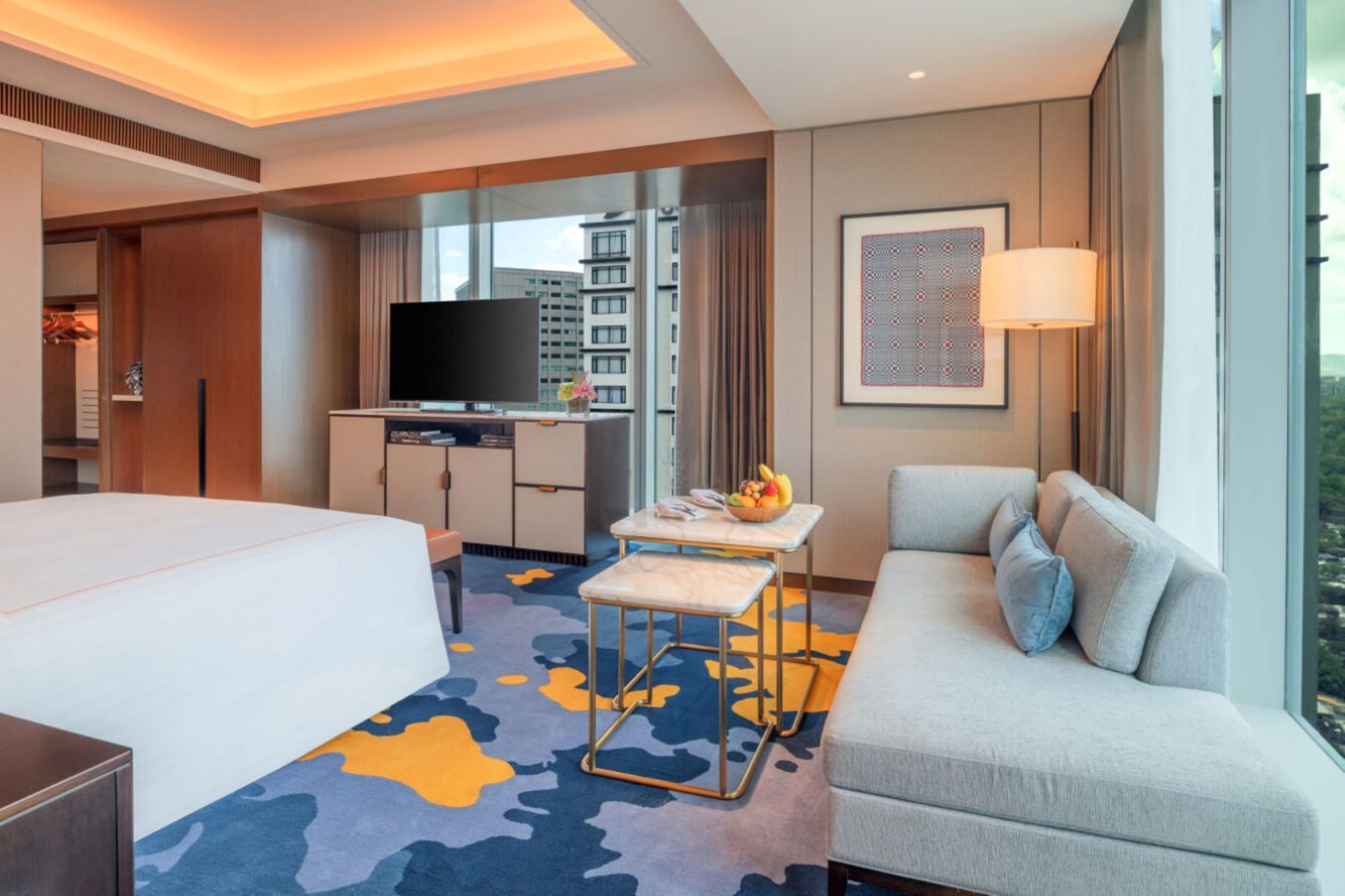 A hotel room with a bed, a sofa, a small table with fruit, a TV, and a city view.