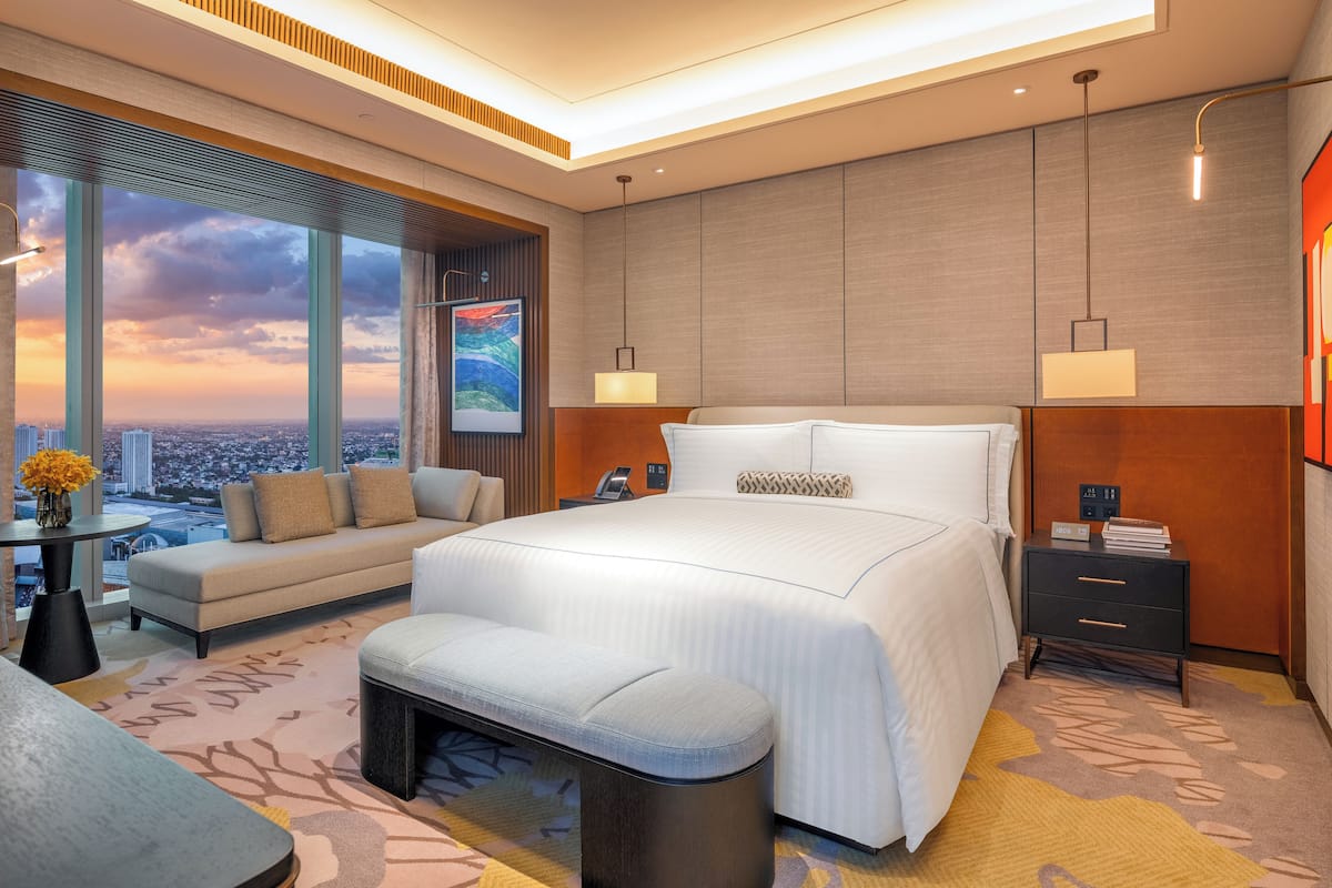A modern hotel room with a large bed, a sofa, a bench, a nightstand, and a view of the cityscape.