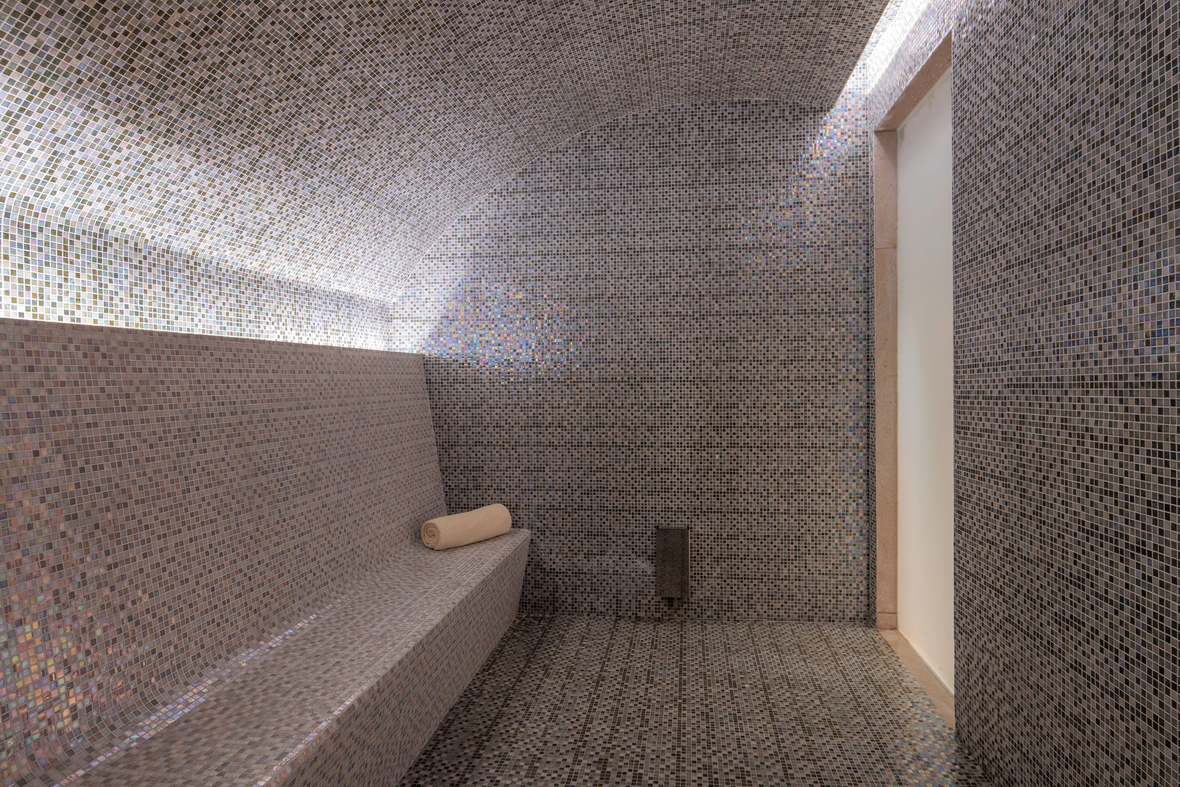 A room with mosaic tile walls and floor, a bench with a towel, and a small window.