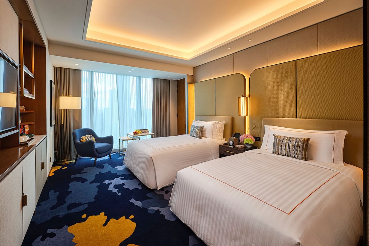 A hotel room with two beds, a desk, a chair, a TV, and a large window with curtains.