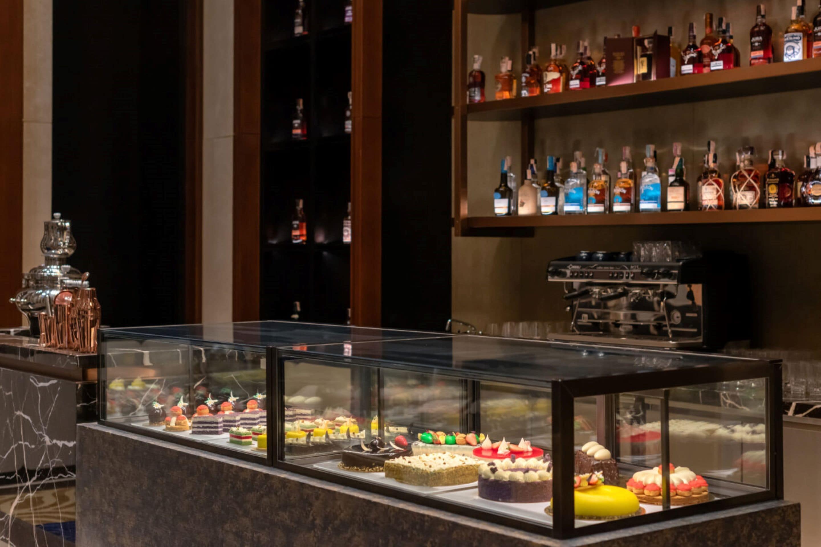 A dessert display case with various cakes and pastries, a bar area with shelves of liquor bottles, and marble flooring.