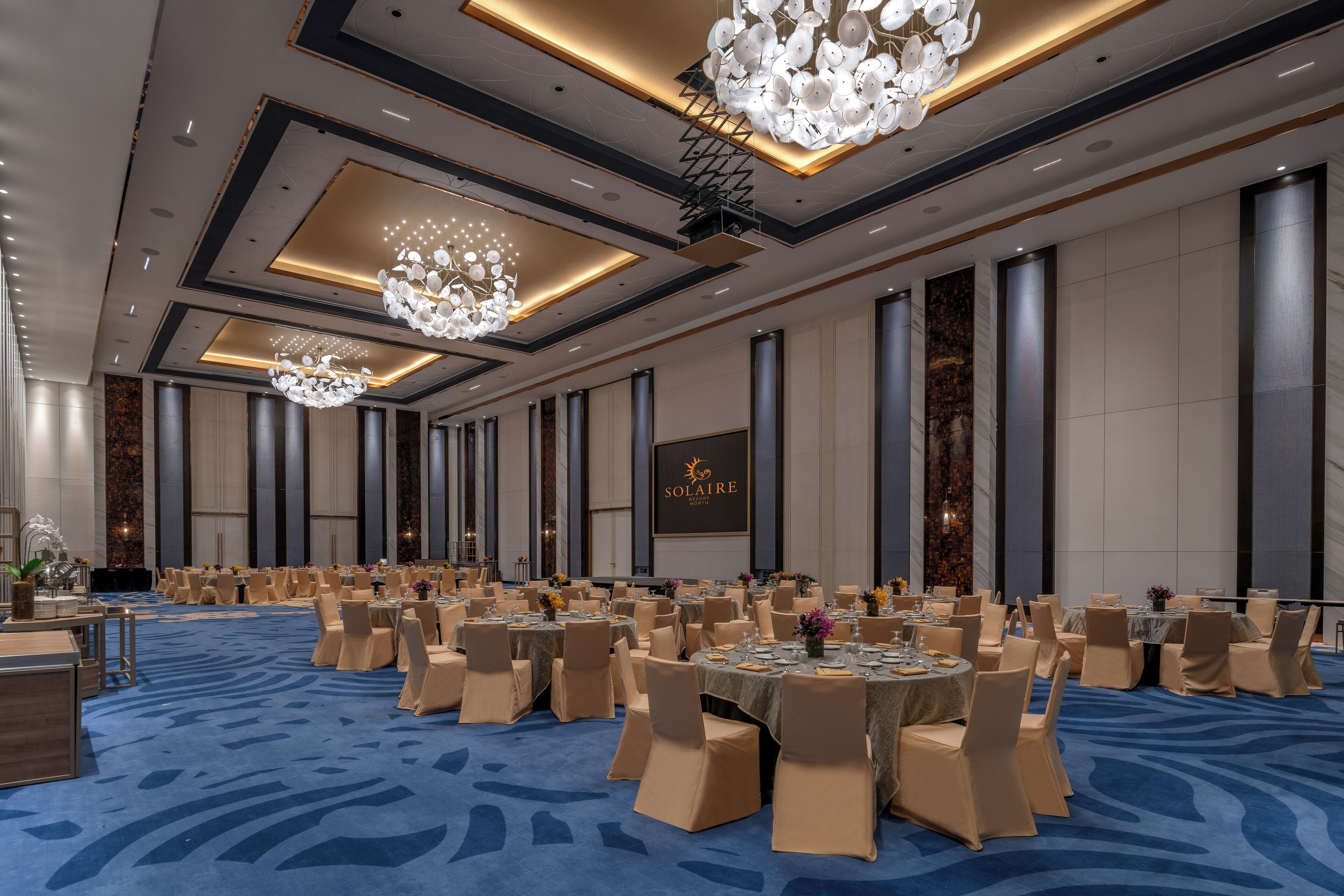 A large, well-lit conference room with round tables, beige chairs, and a blue carpet.