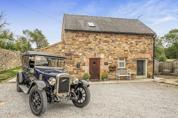 A vintage car parked in front of a stone building with a wooden door and a small garden area with a bench and table.