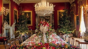 Un comedor amplio y elegante con una larga mesa dispuesta para una comida, flanqueado por dos árboles de Navidad y rodeado de paredes rojas y candelabros.