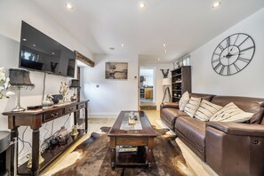 Un salon avec un canapé en cuir marron, une table basse en bois, un téléviseur fixé au mur et une grande horloge murale.