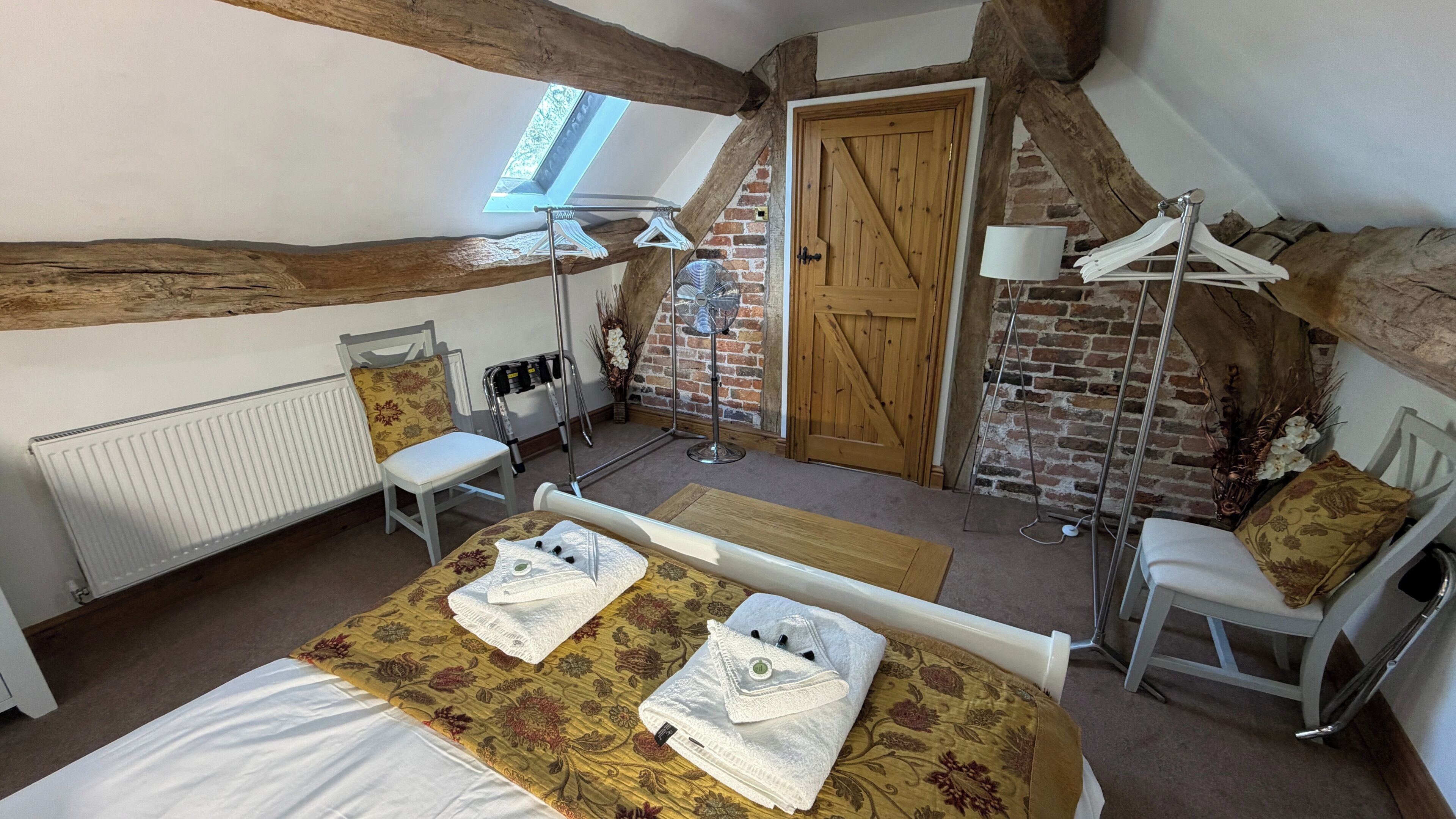 Een kamer met een schuine kap, een bed met handdoeken, een stoel, een radiator, een ventilator en een deur.