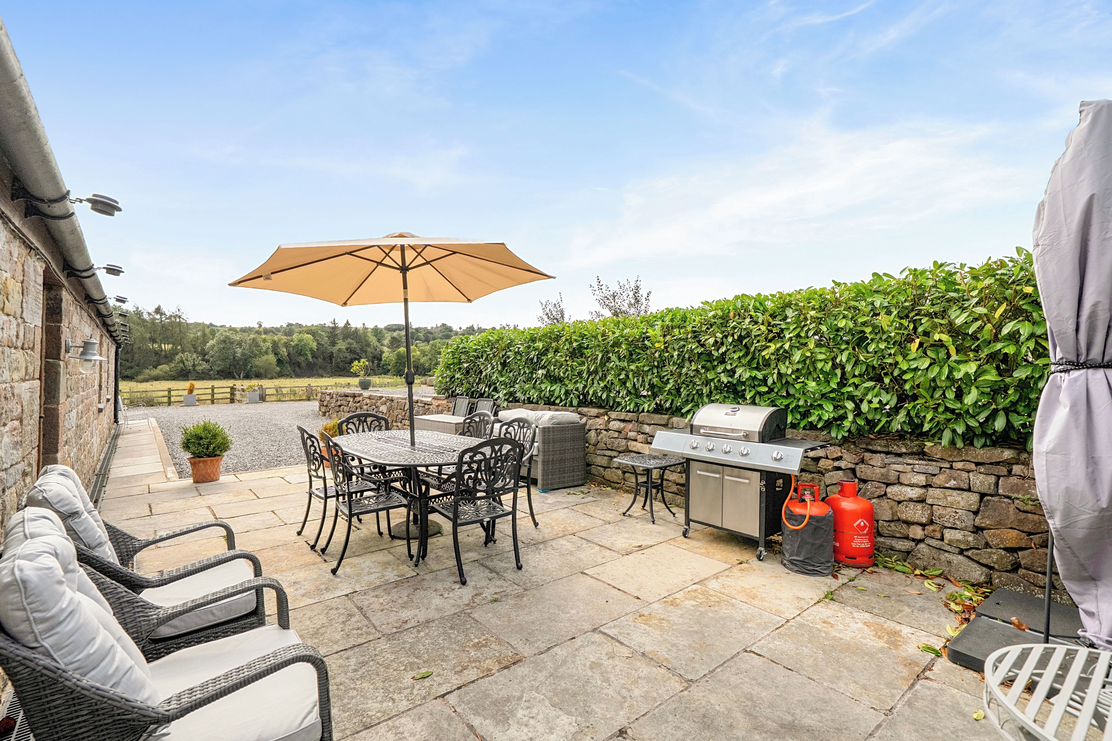 An outdoor patio with a table and chairs, a grill, and a hedge.