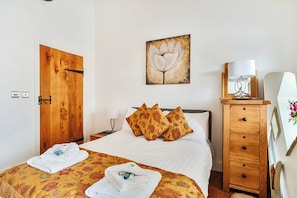 A hotel room with a wooden door, a bed with floral bedding, a nightstand with a lamp, a wooden dresser, and a framed artwork on the wall.