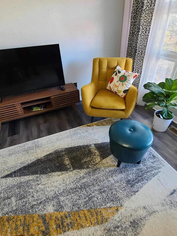Ein Wohnzimmer mit einem gelben Sessel, einem blauen Hocker, einem Fernseher auf einem Holzboden und einem großen Fenster mit gemusterten Vorhängen.