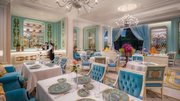A luxurious dining room with ornate chandeliers, blue upholstered chairs, and a bar area with a bartender.