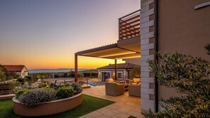 Um alpendre coberto com assentos, piscina e vista para o pôr do sol.