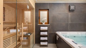 Uma sala de spa moderna com sauna de madeira, área de chuveiro com paredes de vidro, mesa com tampo de vidro e toalhas, prateleira na parede com uma orquídea em vaso e uma grande banheira de hidromassagem.