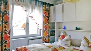 A bedroom with a bed, a window with curtains, a radiator, and a shelf with decorative items.