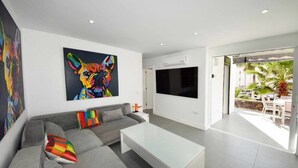 Una sala de estar moderna con un sofá gris, almohadas coloridas, una mesa de centro blanca, un televisor grande en la pared y vistas al exterior.