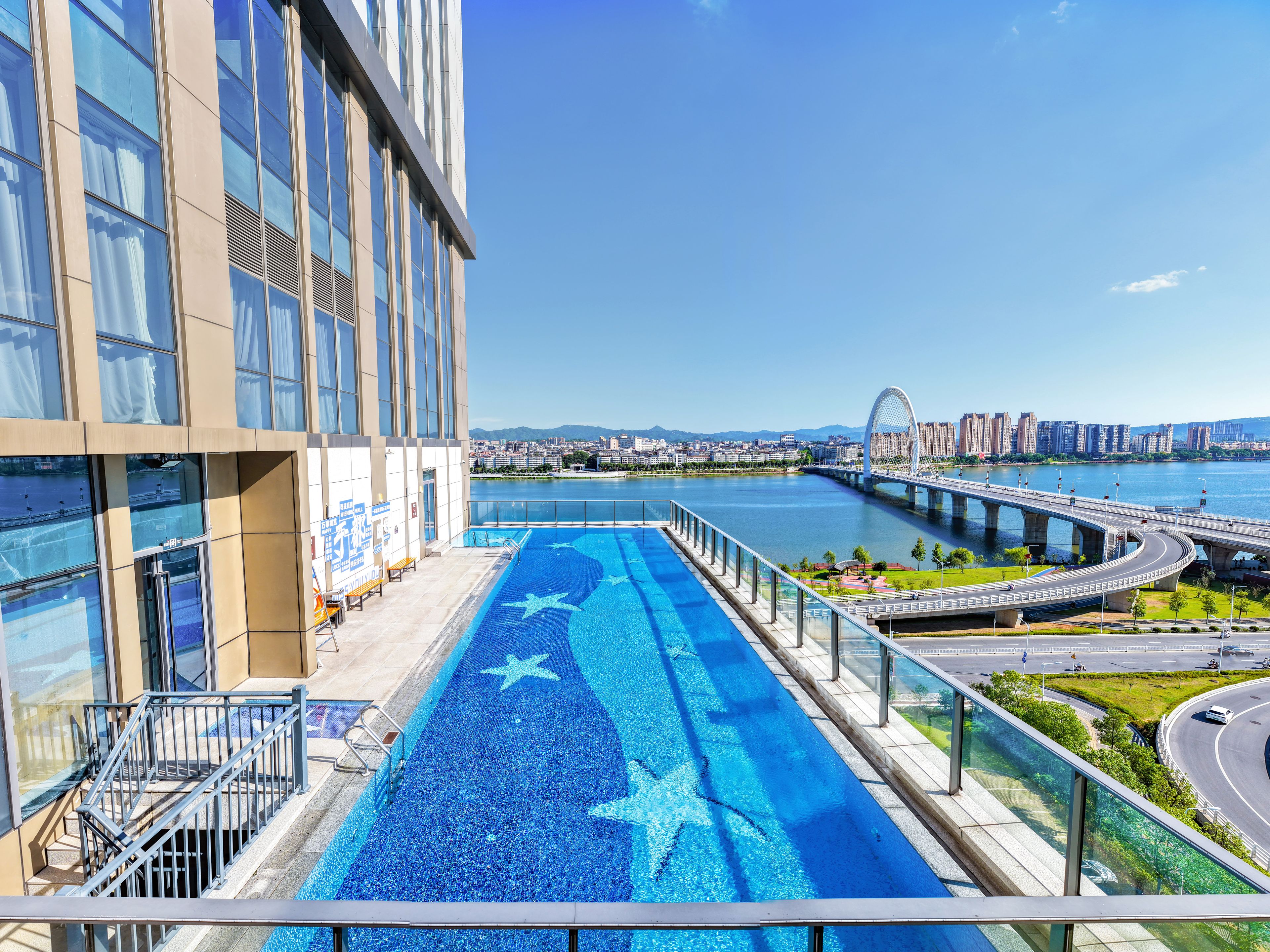 Edificio moderno con piscina en la azotea que presenta un diseño de estrellas, vista de un río y un puente al fondo.