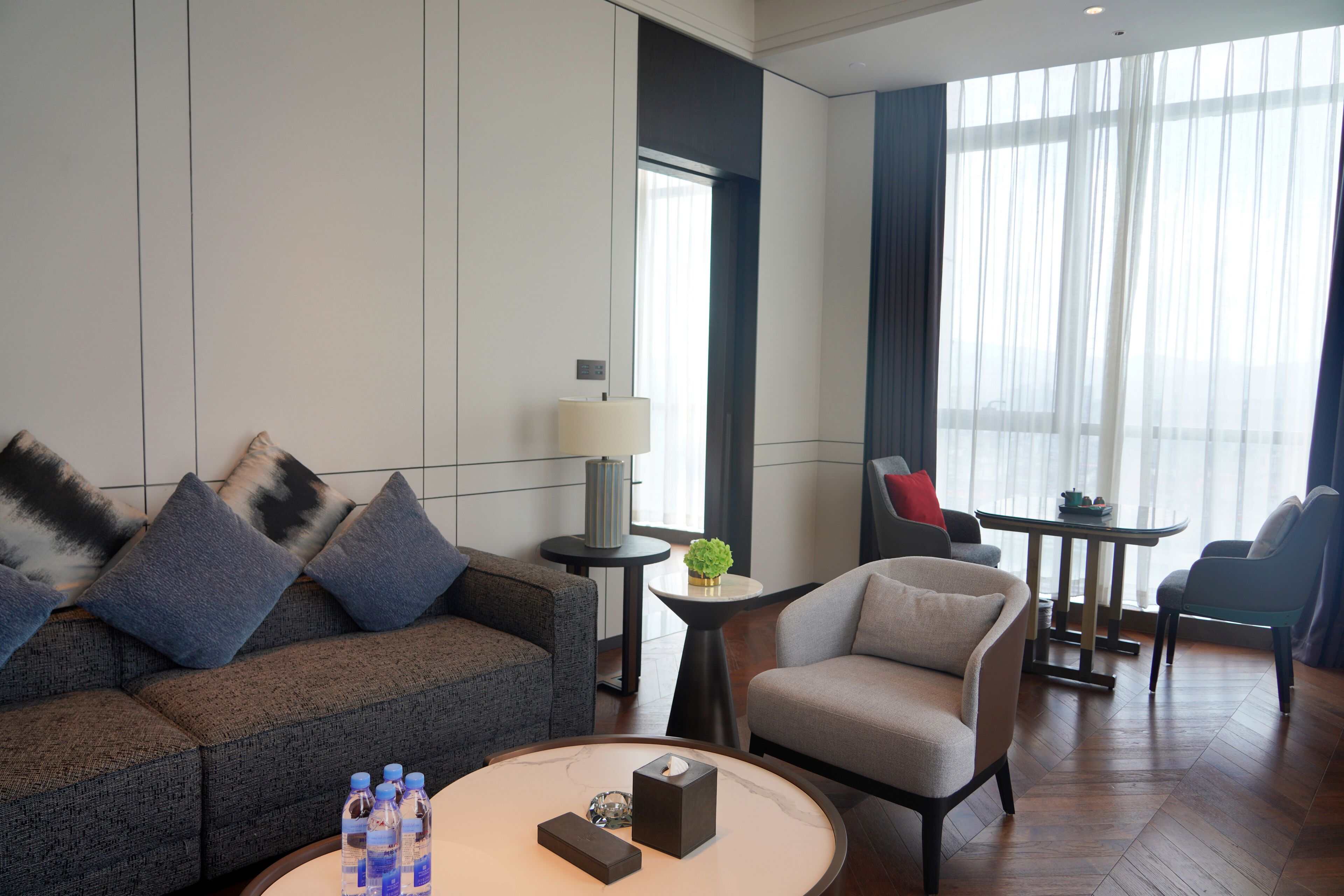 A modern hotel room with a sofa, armchair, round coffee table, and a small side table with a lamp and decorative items.