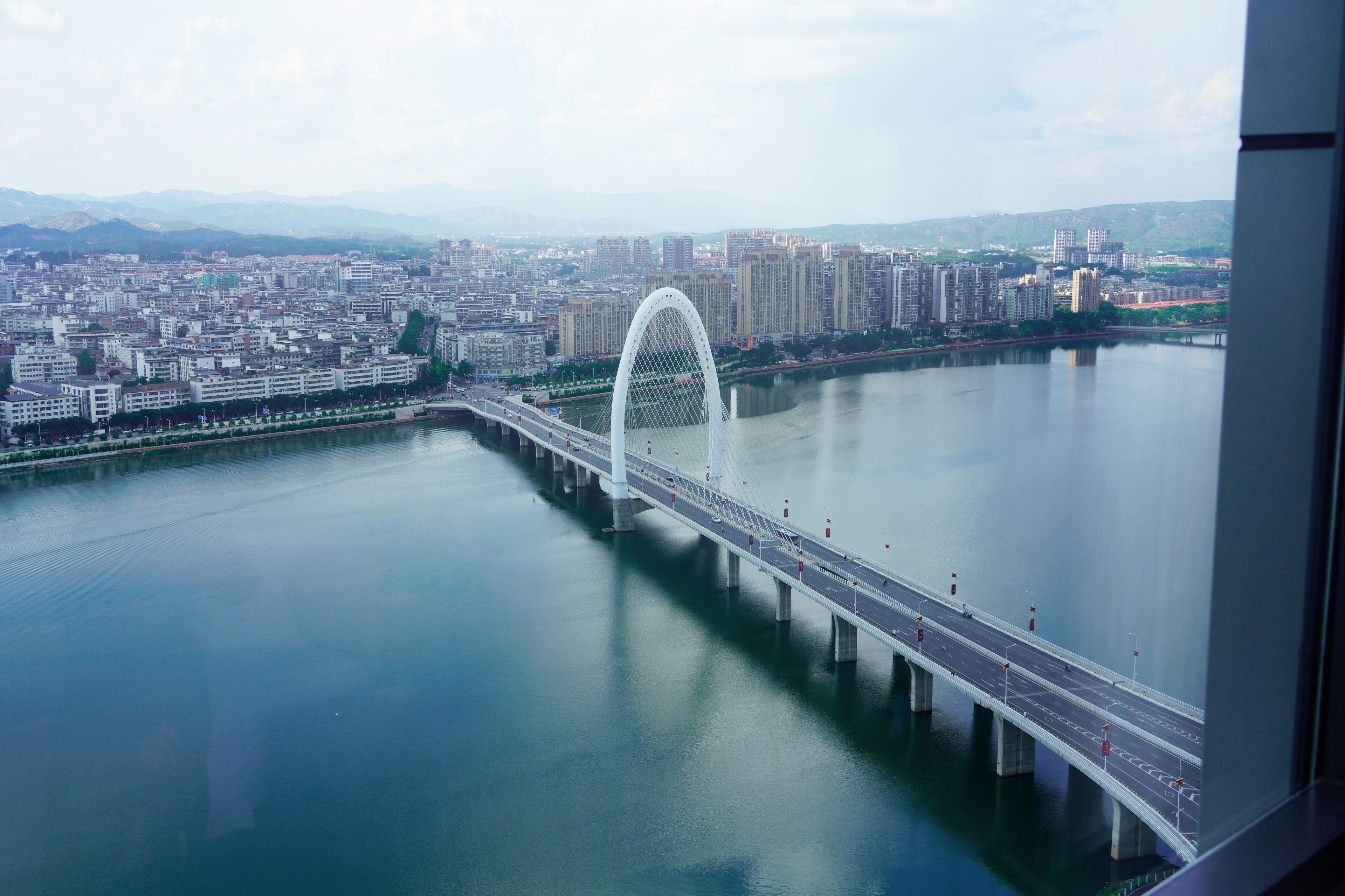 A modern bridge spanning a calm river, surrounded by a cityscape with high-rise buildings and a distant mountain range.