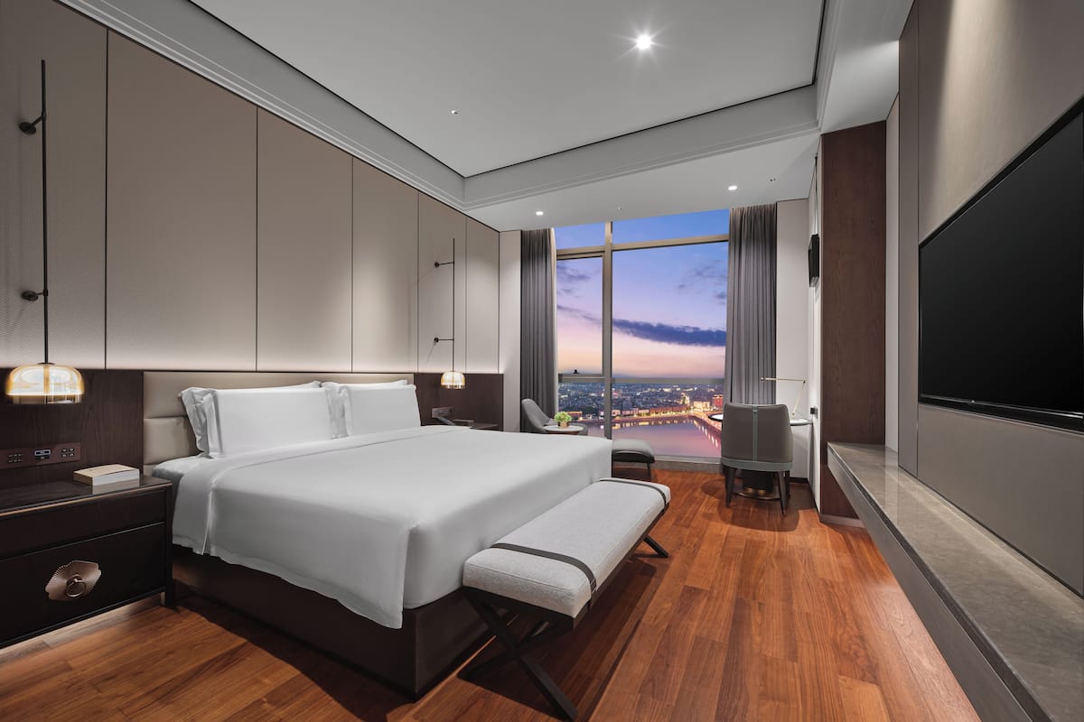 Une chambre d’hôtel moderne dotée d’un grand lit, de tables de chevet, d’un banc, d’une télévision et offrant une vue sur le paysage urbain.