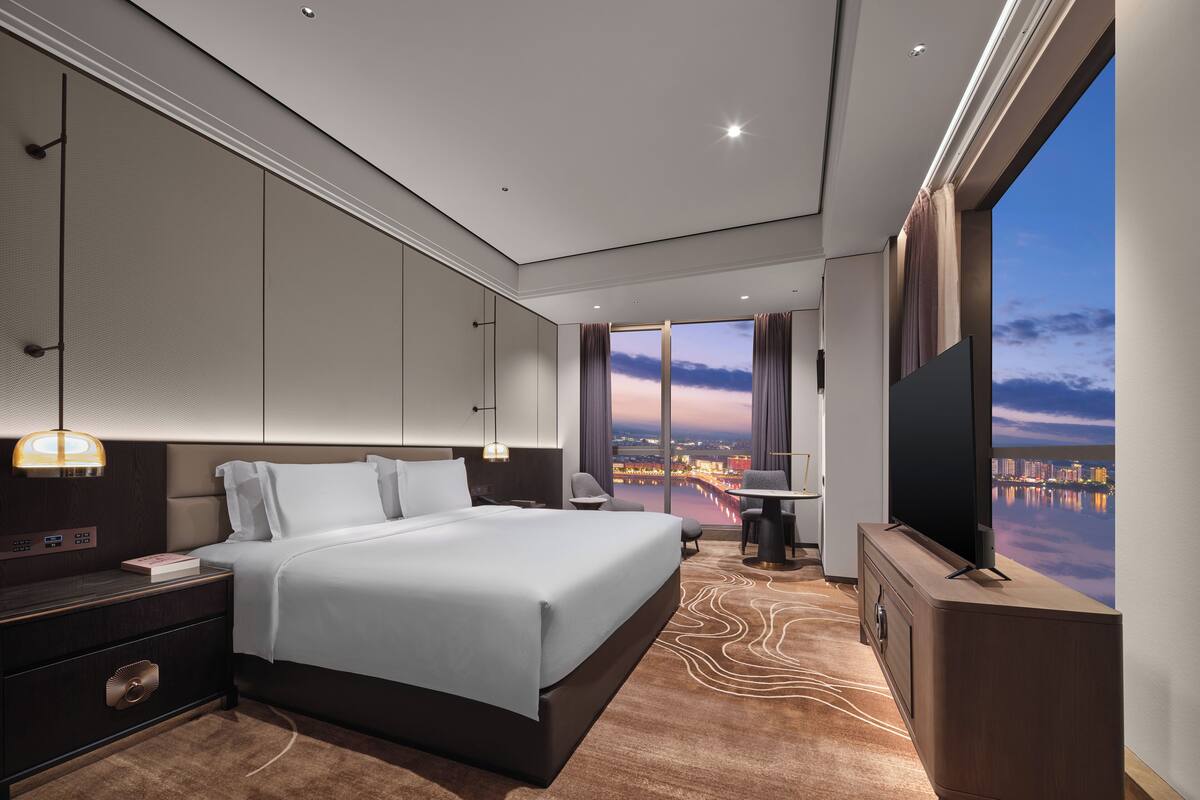 Une chambre d’hôtel moderne dotée d’un grand lit, d’un bureau, d’une télévision et offrant une vue sur le paysage urbain au crépuscule.
