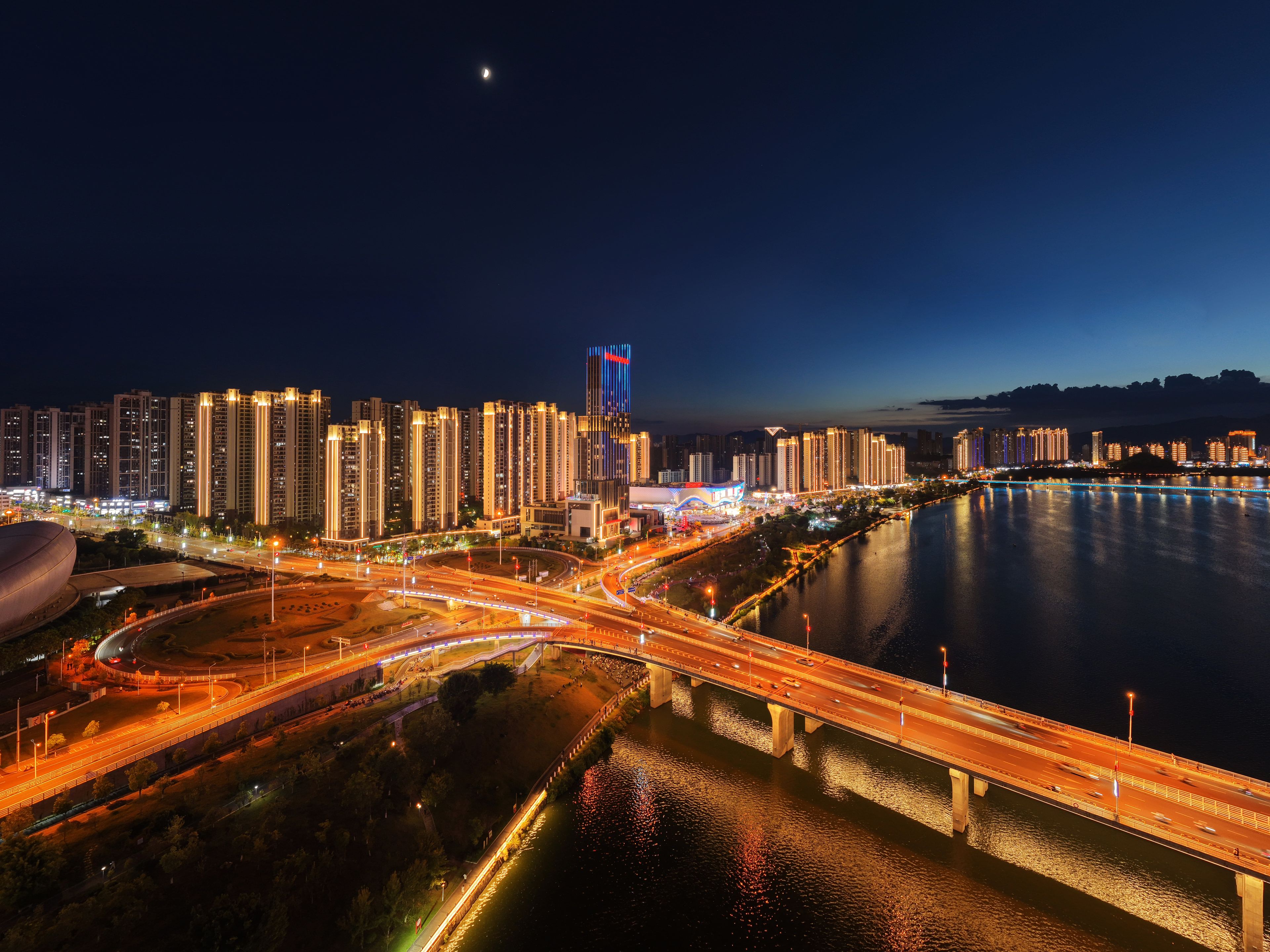 Une vue nocturne d’une ville, avec des bâtiments illuminés, une rivière et un pont.