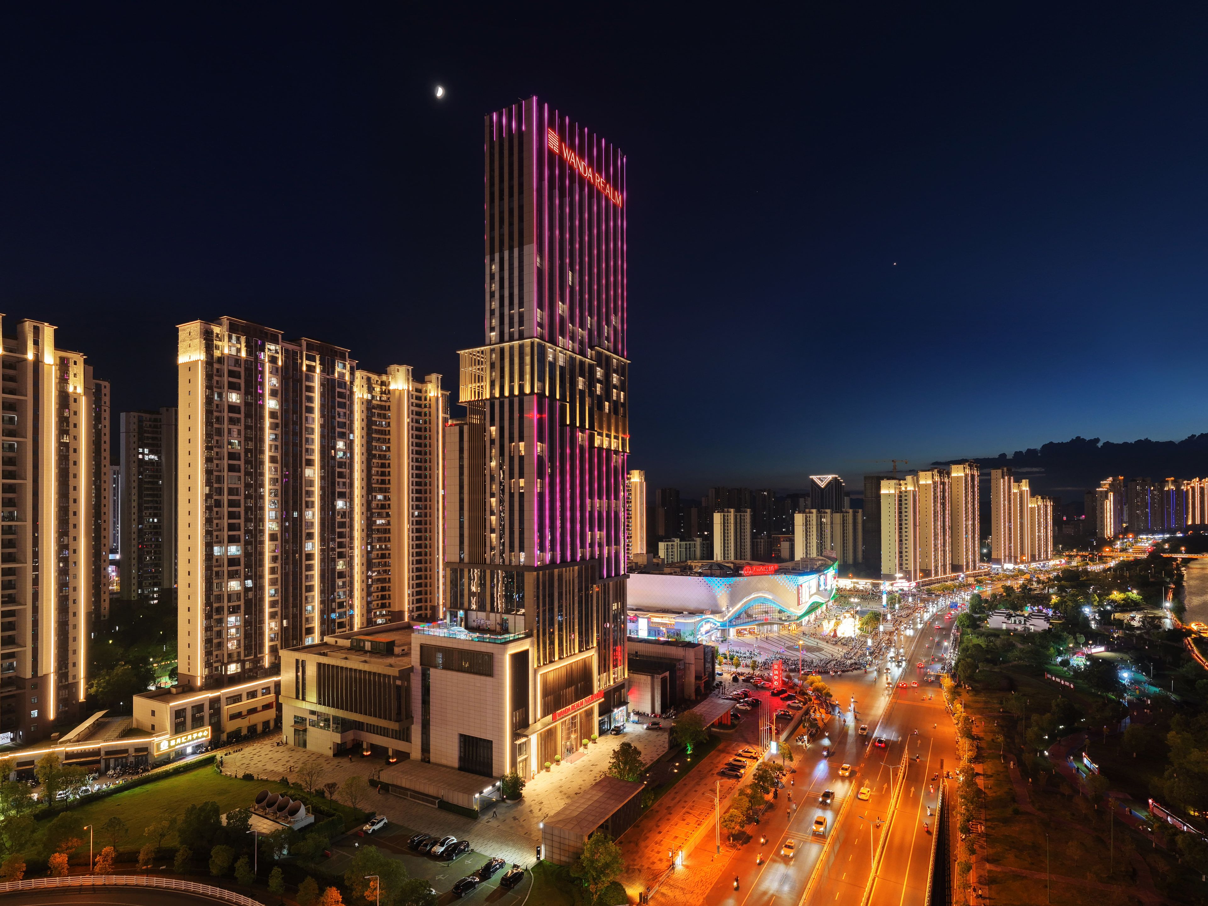 A high-rise building with illuminated signage, surrounded by other tall buildings and a busy street with vehicles.