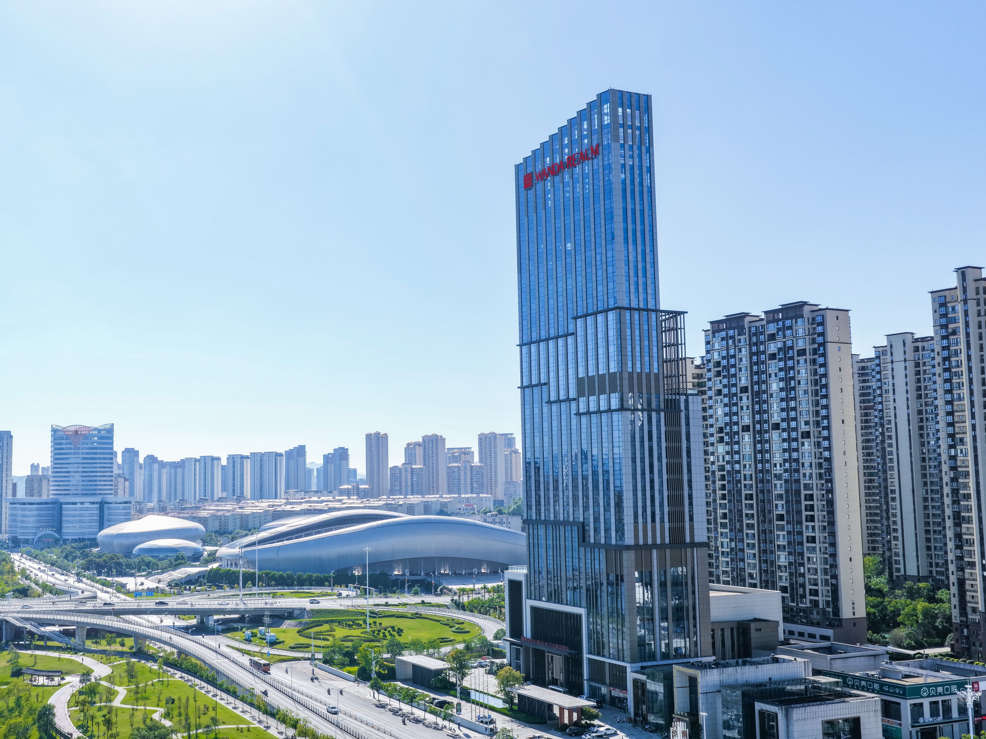 A modern high-rise building with a distinctive red logo, surrounded by other high-rise buildings and a green park area.