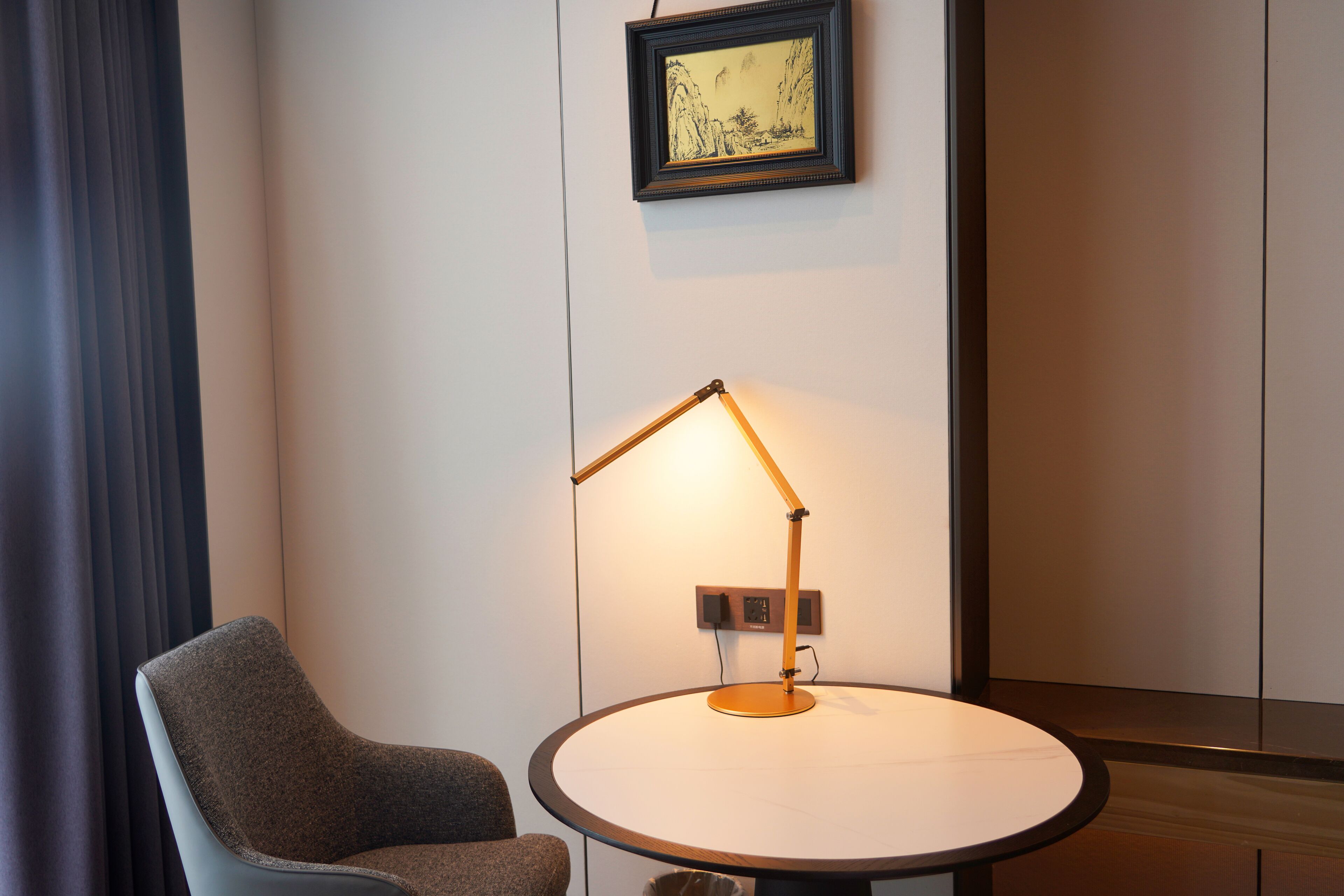 A round table with a lamp, a chair, and a framed picture on the wall.