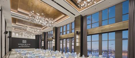 A spacious banquet hall with round tables and chairs, large windows offering a city view, and a modern chandelier.