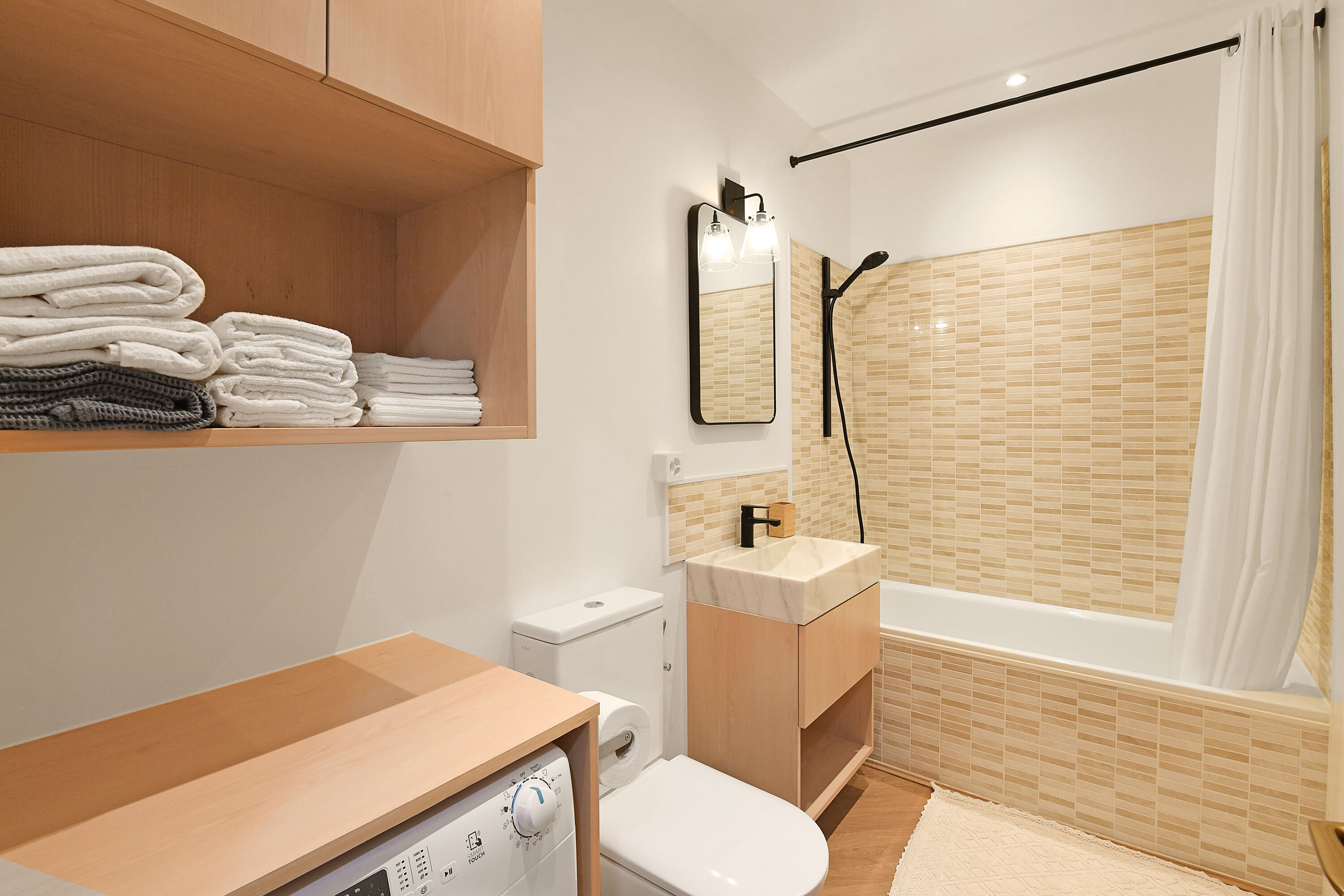 A modern bathroom with a wooden vanity, a toilet, a sink with a mirror, and a shower with beige tiles.