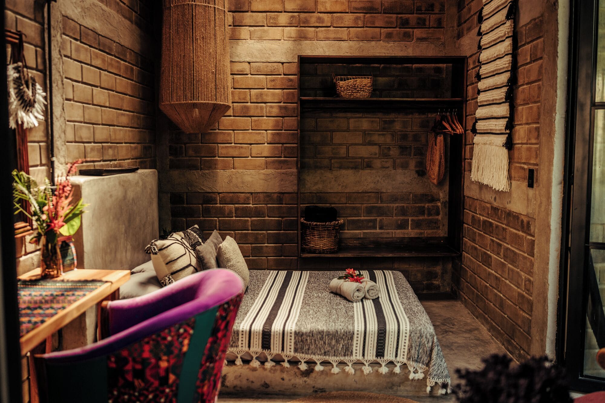 A cozy room with a brick fireplace, a cushioned bench, a woven basket, and a small table with a vase.